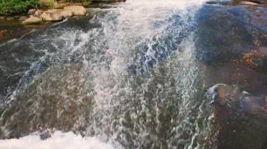 Waterfall Over Low Rocks Background 4K Loop features an aerial shot of water flowing over large boulders/rocks creating an abstract pattern in a loop.