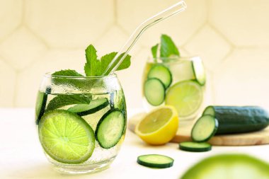 Masada saman olan bardaklarda soğuk, taze yaz içkisi. Dilimlenmiş limonata, limon, salatalık, nane ve buzlu parlak sarı zemin. Kapatın. Seçici odaklanma. Bulanık arkaplan.