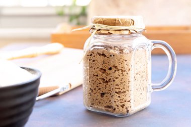 Cam kavanozda aktif ekşi ekmek açıcı yakın plan. Çavdar ekmeği ve mutfak gereçleri için kırsal arka planda bırakılır. Geleneksel ekmek pişirme konsepti. Seçici odaklanma. Bulanık arkaplan.