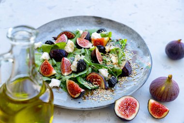  İncir, beyaz peynir, roka ve gri seramik tabakta susam tohumlarıyla tatlandırılmış siyah zeytinli taze sağlıklı yaz salatası ve ön planda bulanık cam zeytinyağı şişesi. Yakın plan. Seçici odaklanma. Bulanık arkaplan.