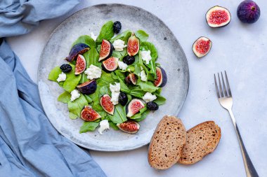 Taze sağlıklı karışık yaz salatası incir, beyaz peynir, roka ve gri seramik tabakta siyah zeytin, iki parça tam buğday ekmeği ve gri arka planda gri bez. Yukarıdan aşağıya doğru. Yakın plan..