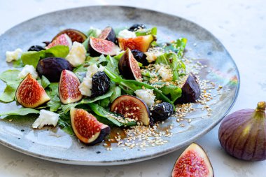 Yakın plan sağlıklı karışık yaz salatası incir, beyaz peynir, roka ve gri arkaplandaki gri seramik tabakta susam tohumlu siyah zeytin. Seçici odaklanma. Bulanık arkaplan.