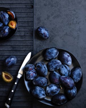 Taze sulu mavi bütünün üst görüntüsü ve siyah tabakta erikler ve koyu gri arka planda mutfak bıçağı. Gösterişsiz fotoğraf. Yakın plan. Düz kompozisyon. Boşluğu kopyala.