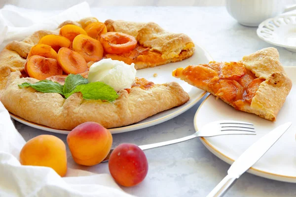 Close-up of traditional French galette with apricots, mint and cream with cut piece on white plate, fork and knife on white marble table. Summer baking. Selective focus. Blurred background.