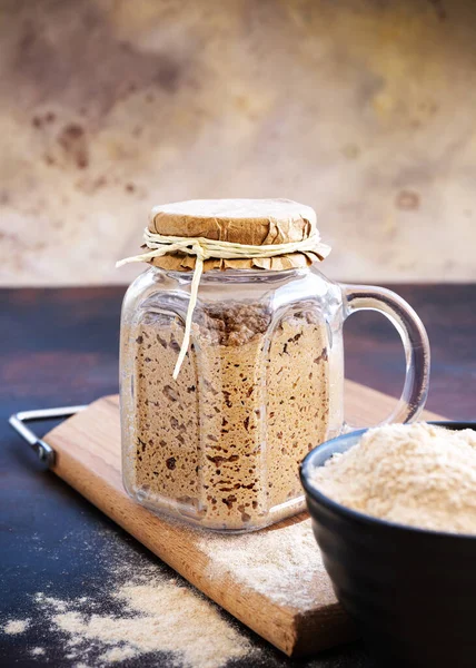 Vertical shot of active sourdough starter in glass jar. Rye leaven for bread and cup of flour on wooden cutting board on black and brown rustic background. Traditional bread baking concept. Close-up. Selective focus. Blurred background.