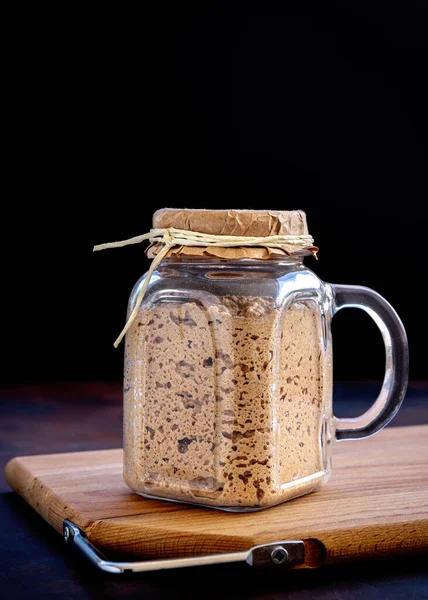 Active sourdough starter in glass jar. Rye leaven for bread on wooden cutting board on black background. Close-up. Dark low key photo. Selective focus. Traditional bread baking concept.