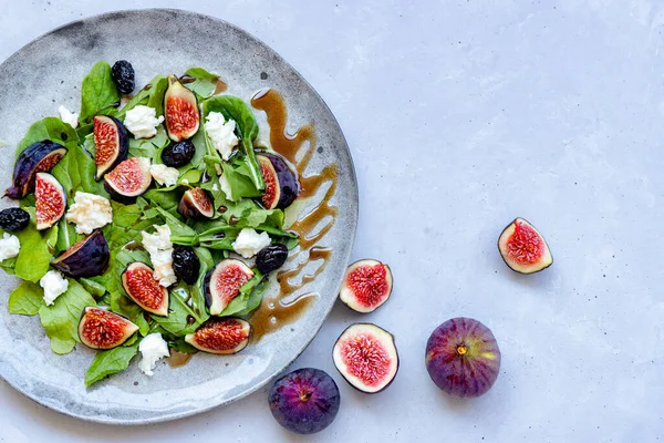 Top view of fresh healthy mixed summer salad with figs, white cheese, arugula and black olives seasoned with sauce on grey ceramic plate on grey background. Close-up. Flat lay composition. Copy space.