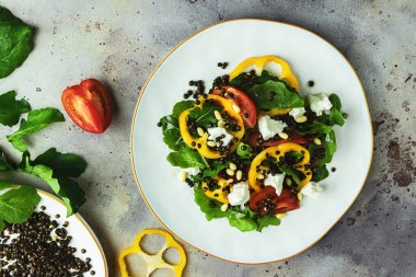 Top down view of fresh healthy mixed vegetable salad with black lentil, cheese, yellow bulgarian pepper, arugula and pine nuts on white ceramic plate on rustic gray background. Top view. Close-up. Flat lay composition.