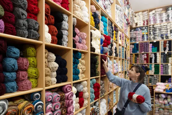 Beautiful smiling caucasian woman choosing yarn in knitting shop. Variety of colorful yarn balls in craft store. Hobby handmade concept. Selective focus.