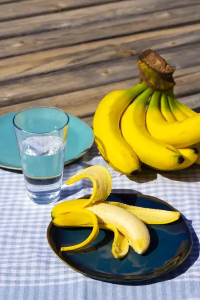 黄色い熟したバナナ プレートにバナナと青いテーブルクロスの水のグラス 日当たりの良い木の表面に新鮮な有機果物 まだ人生の写真 選択的な焦点 バックグラウンド — ストック写真