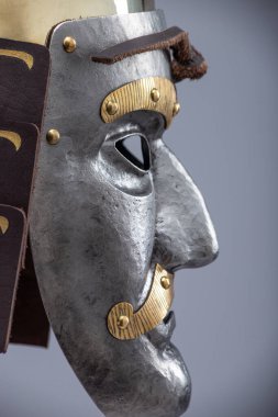 Tarihi metalik savaş miğferi ve entegre yüz maskesi. Detaylı metalik bir yüz maskesi, göz delikleri, dekoratif elementler ve yan flapları olan sivri uçlu çelik miğfer..