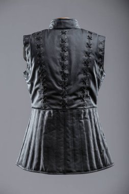 Dantelli ve kalçalı siyah deri kolsuz ceket. Bu resim siyah, kolsuz bir ceket ya da yüksek yakalı bir yelek gösteriyor. Sergide dekoratif süsleme ayrıntıları ve önünde gümüş tokalar bulunan ayarlanabilir kayışlar yer alıyor..