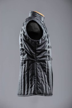 Dantelli ve kalçalı siyah deri kolsuz ceket. Bu resim siyah, kolsuz bir ceket ya da yüksek yakalı bir yelek gösteriyor. Sergide dekoratif süsleme ayrıntıları ve önünde gümüş tokalar bulunan ayarlanabilir kayışlar yer alıyor..