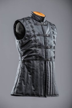 Dantelli ve kalçalı siyah deri kolsuz ceket. Bu resim siyah, kolsuz bir ceket ya da yüksek yakalı bir yelek gösteriyor. Sergide dekoratif süsleme ayrıntıları ve önünde gümüş tokalar bulunan ayarlanabilir kayışlar yer alıyor..