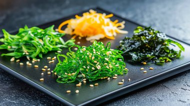 Üç çeşit susam tohumuyla süslenmiş deniz yosunlarını içeren, parlak siyah bir tabakta canlı bir deniz yosunu salatası gösterisi..