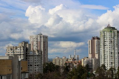 Modern bir şehirde yeşil parkın etrafında boruları olan yüksek binalar ve endüstriyel bir fabrika.