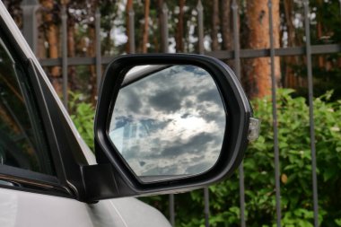 The cloudy sky is reflected in the mirror of a car parked near the iron fence