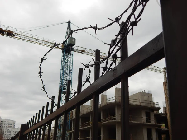 Stopped high-rise construction crane behind a metal fence with barbed wire in focus on foreground