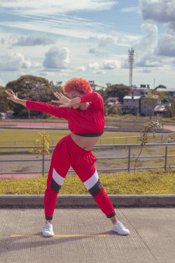 Joven afro de cabello naranja bailando al estilo urbano en parque local pista de atletismo, vejetaryen y cielo hermosos de fondo