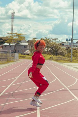 Joven afro de cabello naranja bailando al estilo urbano en parque local en la pista de atletismo, vejetaryen y cielo hermosos de fondo
