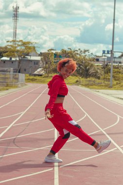 Joven afro de cabello naranja bailando al estilo urbano en parque local en la pista de atletismo, vejetaryen y cielo hermosos de fondo