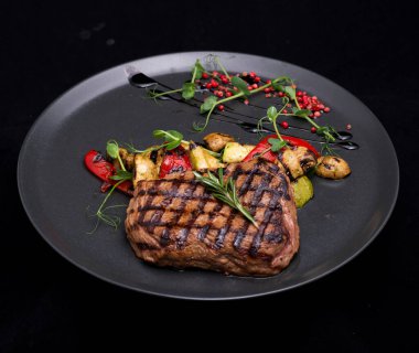 Biftek, ızgara sebze, baharat ve otlar koyu bir arka planda.