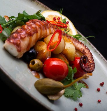 Izgara ahtapot ve siyah tabakta bitkilerle birlikte sebze.