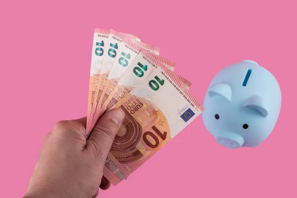 A man\'s hand holds out several banknotes of 10 euros to a piggy bank on a pink background
