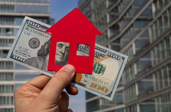 Symbolic paper house and a 100 american dollar bill against the backdrop of a modern high-rise building