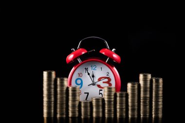 Many columns of coins in denominations of 100 Kazakhstani tenge and a red classic alarm clock on a red background