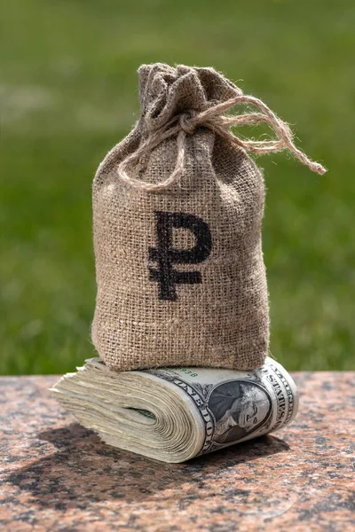A bag for money with the symbol of the Russian ruble stands on a stack of banknotes in denominations of 1 US dollar