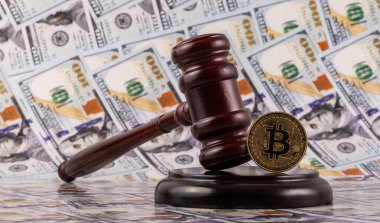 Üzerinde Bitcoin kripto para sembolü olan altın bir sikke ve arka planda 100 dolarlık banknotlar bulunan bir yargıç tokmağı.