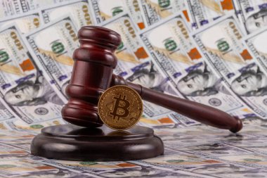 Üzerinde Bitcoin kripto para sembolü olan altın bir sikke ve arka planda 100 dolarlık banknotlar bulunan bir yargıç tokmağı.