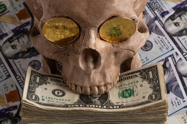 Bir insan kafatasının göz çukurunda Bitcoin kripto para sembolü olan bir altın sikke 100 Amerikan dolarının arka planına karşı.