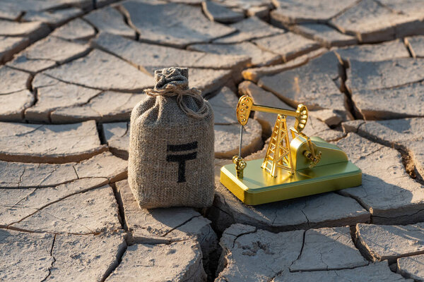 Миниатюрный нефтяной насос и денежный мешок с символом казахстанской валюты тенге на раскаленной глине в пустыне