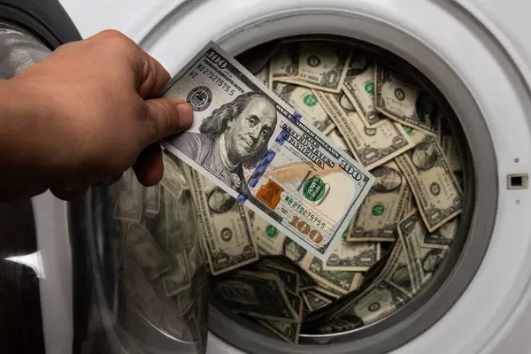 Conceptual story about corruption and money laundering with a washing machine filled with dirty 1 US dollar bills and 100 us dollars in a man\'s hand