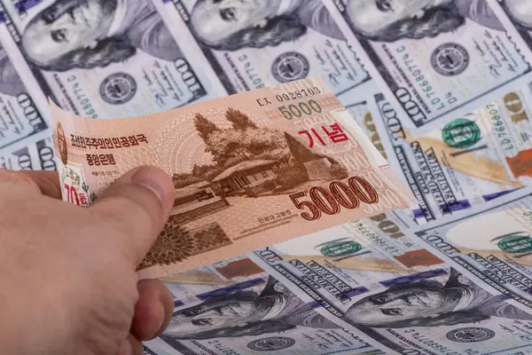 A bill of 5000 North Korean won in a man\'s hand against the background of 100 US dollar bills