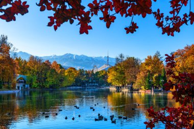 Kazak şehri Almaty 'nin şehir parkındaki bir gölet manzarası karla kaplı dağlar ve bir sonbahar sabahı bir televizyon kulesine karşı.
