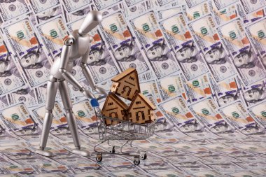 Minyatür bir alışveriş arabasında sembolik ahşap evler ve arka planda 100 dolarlık banknotlarla ahşap bir adam.