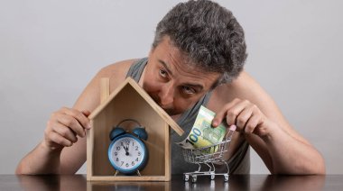 Gri saçlı, orta yaşlı, 100 Euro 'luk banknot, ahşap ev, çalar saat ve süpermarketten alınmış minyatür boş alışveriş arabasıyla yoksullukla ilgili kavramsal bir hikaye.