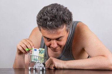 Gri saçlı, orta yaşlı bir adamla yoksulluk hakkında kavramsal bir hikaye. 100 Euro 'luk banknot ve süpermarketten alınmış minyatür alışveriş arabası.