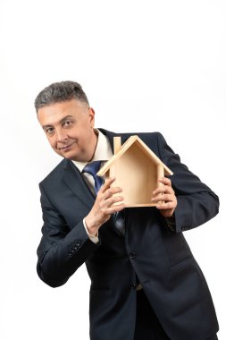Orta yaşlı bir adam beyaz arka planda sembolik ahşap bir ev tutuyor.