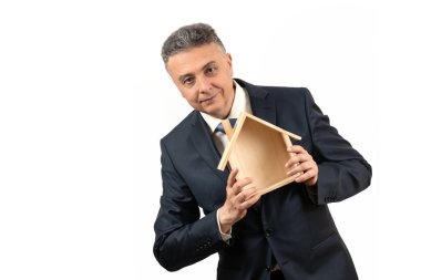 Orta yaşlı bir adam beyaz arka planda sembolik ahşap bir ev tutuyor.
