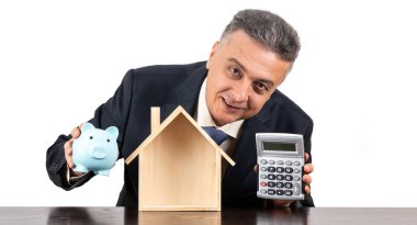 Takım elbiseli orta yaşlı bir adam, mavi kumbara ve sembolik ahşap bir ev.