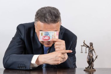 Takım elbiseli bir adam, yüzünün bir kısmını Meksika pezosu ile çiziyor ve Themis 'in figürüne işaret ediyor.