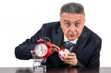 Takım elbiseli orta yaşlı bir adam, alışveriş arabasında kırmızı alarm saati ve beyaz arka planda komik bir kumbara.