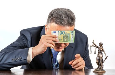 Takım elbiseli orta yaşlı bir adam gözlerini 100 Euro 'luk banknotun ve Themis' in bir figürünün arkasına saklar.