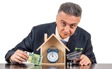 Takım elbiseli orta yaşlı bir adam, 100 euro banknot, sembolik ahşap ev ve beyaz arka planda çalar saat.