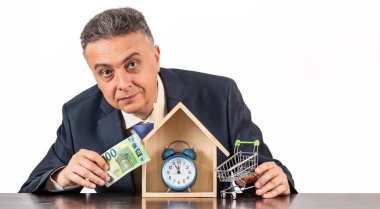 Takım elbiseli orta yaşlı bir adam, 100 euro banknot, sembolik ahşap ev ve beyaz arka planda çalar saat.