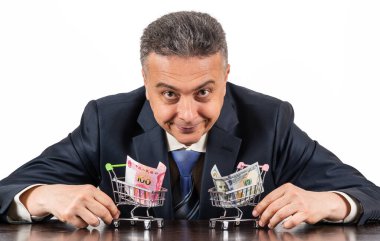 Orta yaşlı takım elbiseli bir adam ve 100 porselen yuan ve beyaz arka planda 100 dolarlık minyatür alışveriş arabaları.